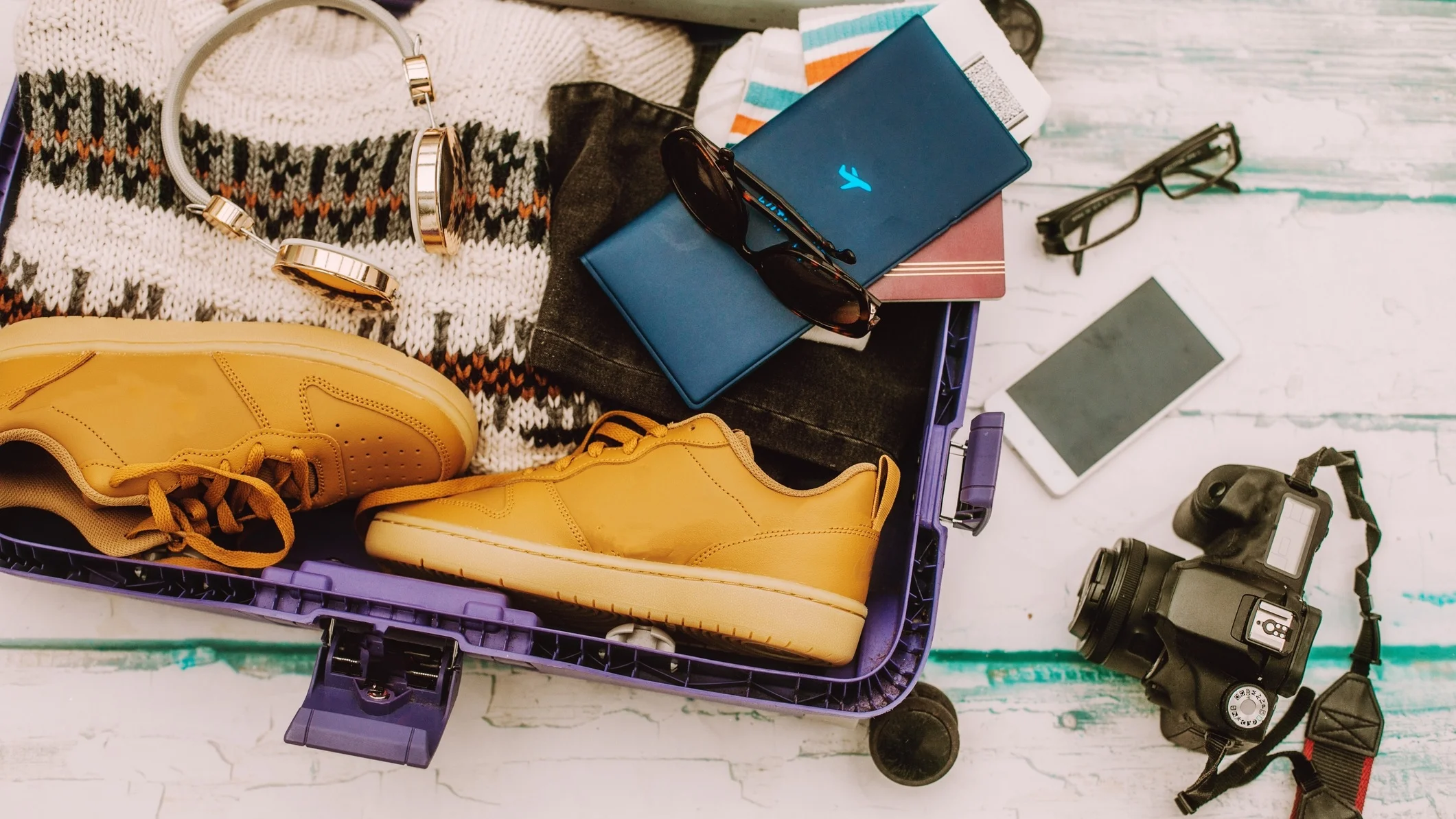 An open suitcase with shoes, a passport, a camera, headphones and sunglass being packed into it.