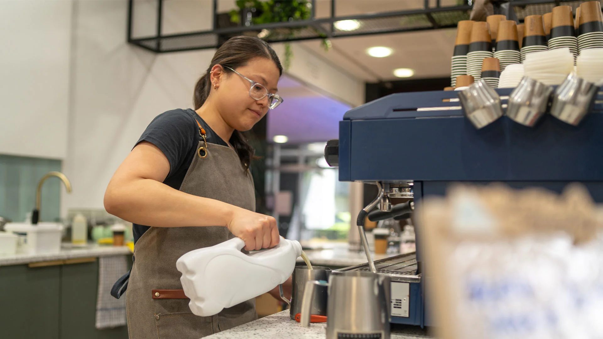 Female barista in NZ pouring milk into jug in order to make a coffee for a customer.