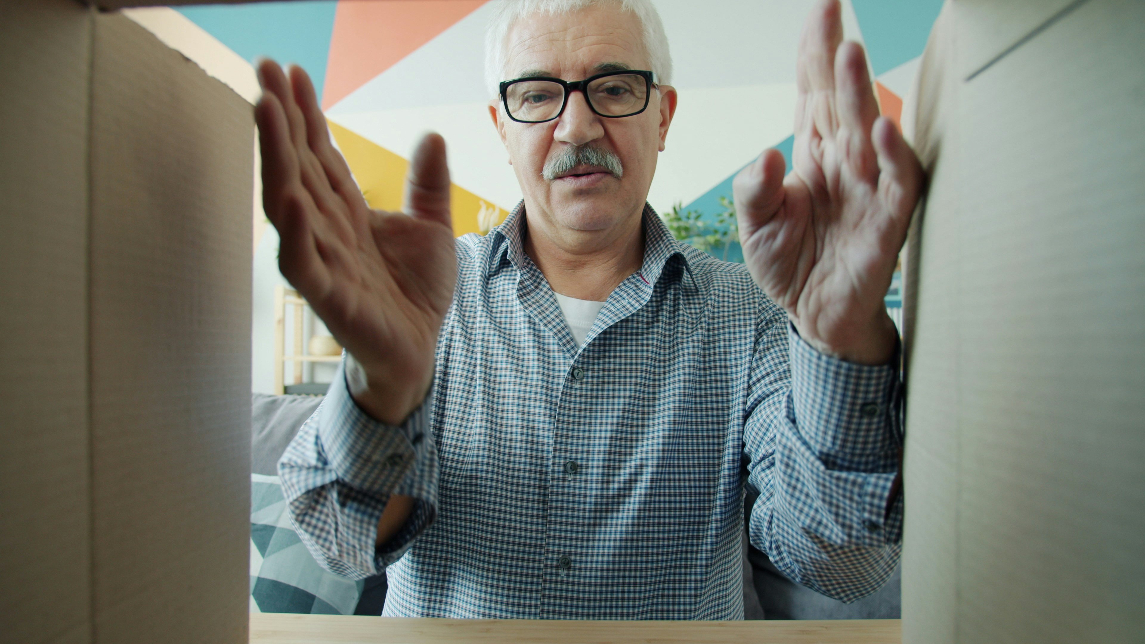 An elderly man looking down into a cardboard box. The perspective is from the bottom of the box looking up at the man. 