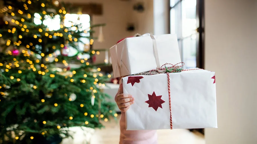 Child holding two big presents that obscure her head, with a Christmas tree with lights behind them.