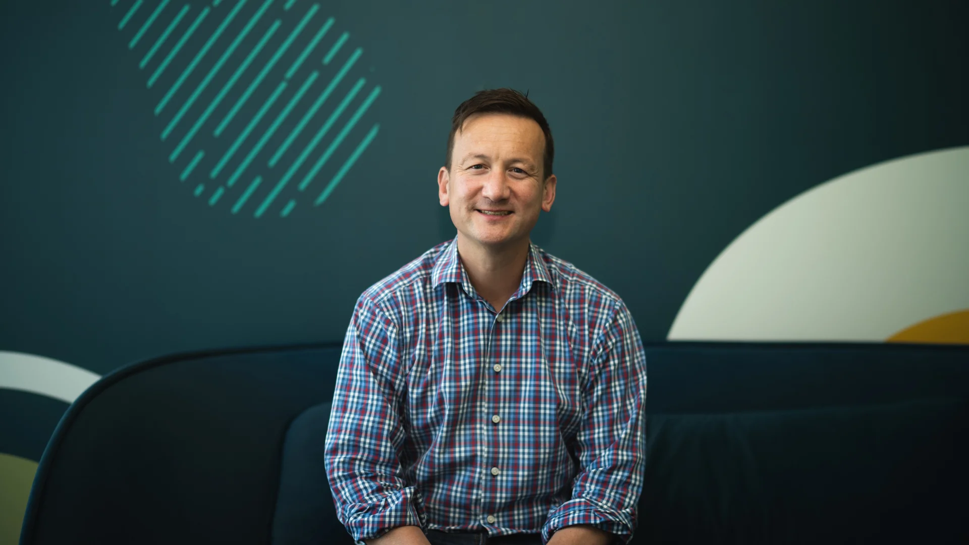 A photo of Alan Clark Trade Me Head of Property. He's sitting on a dark sofa in front of a teal coloured wall.