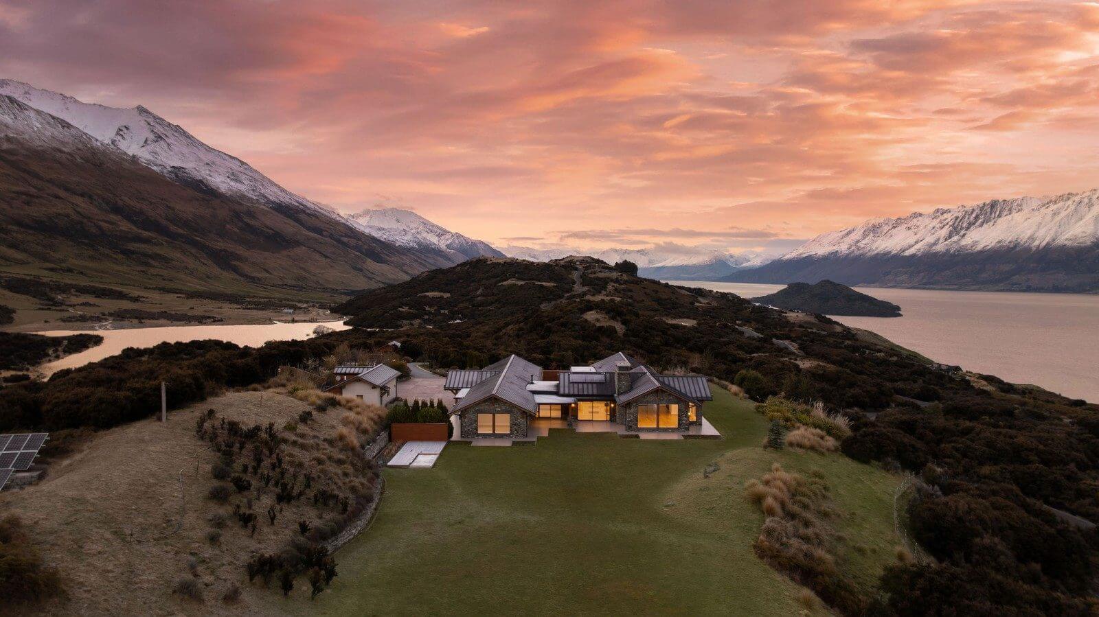 Aerial drone shot of a 650m² architect-designed home at 25 Mount Alfred Ridge, Wyuna, Glenorchy, set on 2.6 hectares with panoramic views across Lake Whakatipu and the Humboldt Ranges at sunset, listed on Trade Me Property.