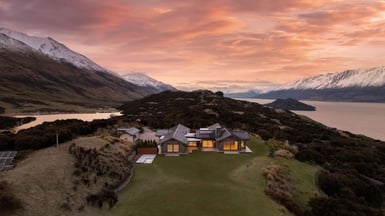Aerial drone shot of a 650m² architect-designed home at 25 Mount Alfred Ridge, Wyuna, Glenorchy, set on 2.6 hectares with panoramic views across Lake Whakatipu and the Humboldt Ranges at sunset, listed on Trade Me Property.