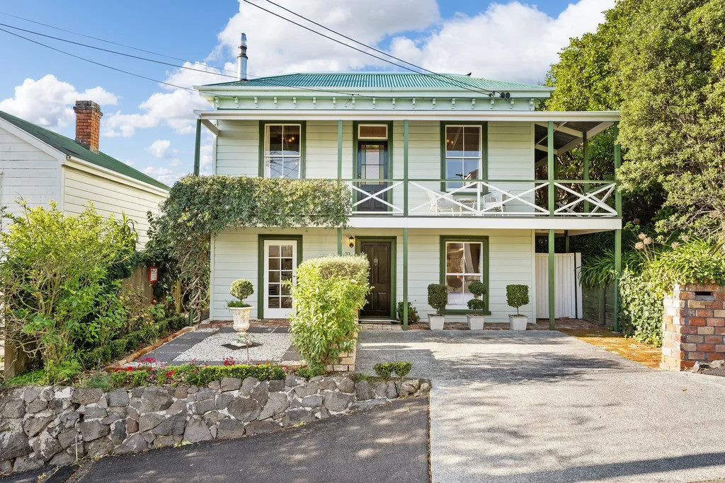 A two-story light green house with a green roof, front balcony, and neatly landscaped garden