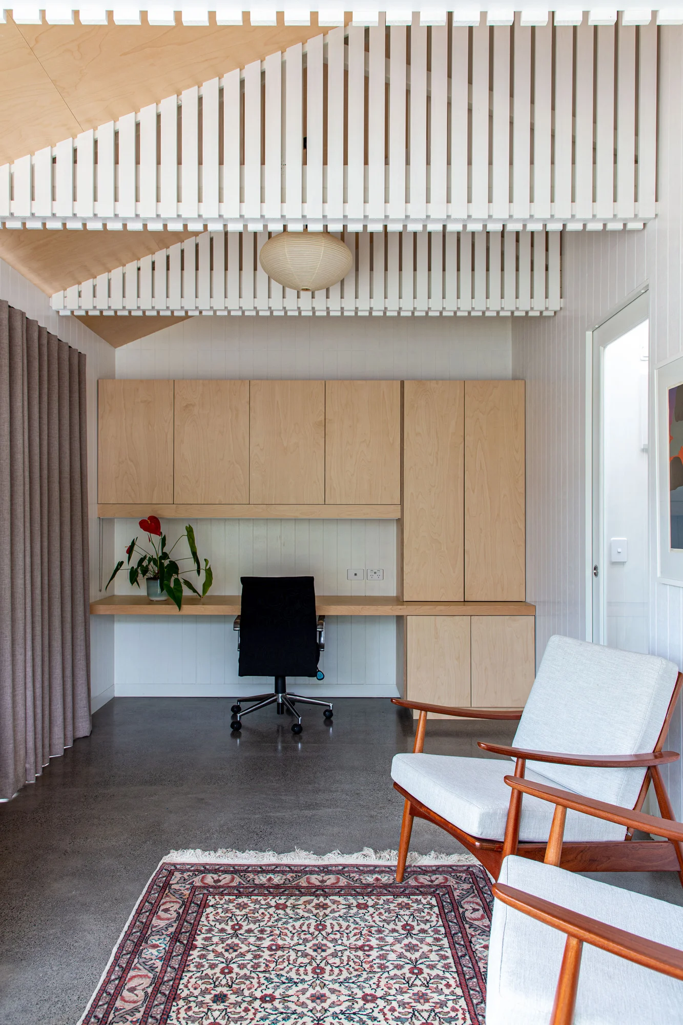 A minimalist office space with concrete floors and wooden cupboards. A red rug in the centre of the floor with two chairs for relaxing around it. On the back wall is a inbuilt desk space which has been built in a light wood.