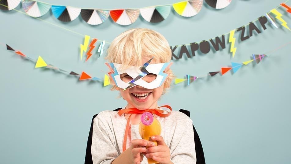 A blonde child in a superhero costume pointing a bubble gun at the camera