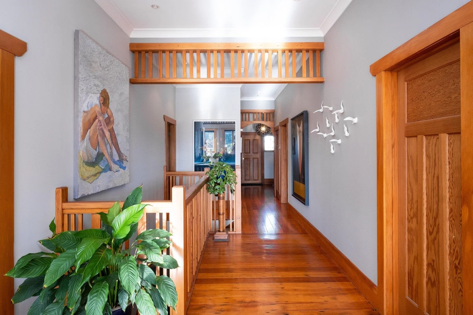 View of hallway with wooden floor and doors. Two house plants and a large painting on the left of a boy at the beach.