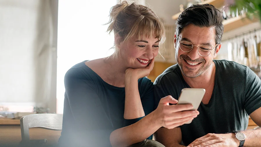 Middle aged couple smiling in their kitchen as the woman looks at her refurbished Samsung phone.