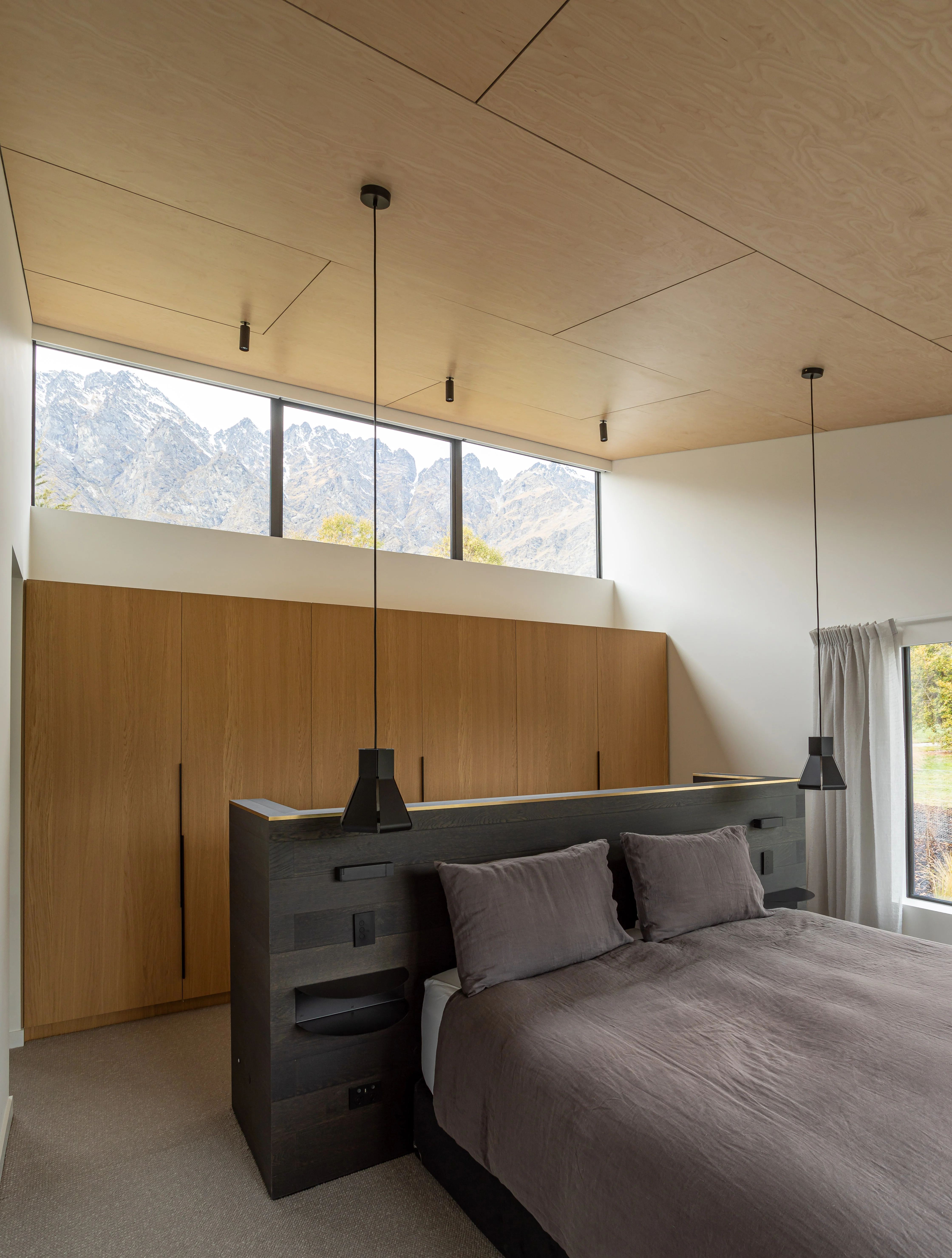 A modern, cozy bedroom. The room features a large bed with grey bedding and multiple pillows. Behind the bed, there is a black half wall acting as a headboard and side tables. Behind this is a wall-to-wall light timber wardrobe. Two black pendant lights hang from the ceiling, providing illumination to the room. A wide window above the wardrobe reveals a scenic view of mountains outside, adding a touch of nature to the room. The walls are painted in a neutral tone, and there’s minimal decoration, giving the room a clean and spacious look.