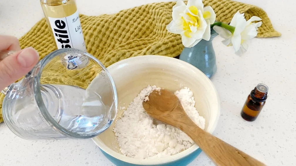 A water from a glass measuring jug into a small pottery bowl filled with baking soda with a small bottle of essential oil, a bunch of daffodils, a bottle of castile soap and a yellow tea towel in the background