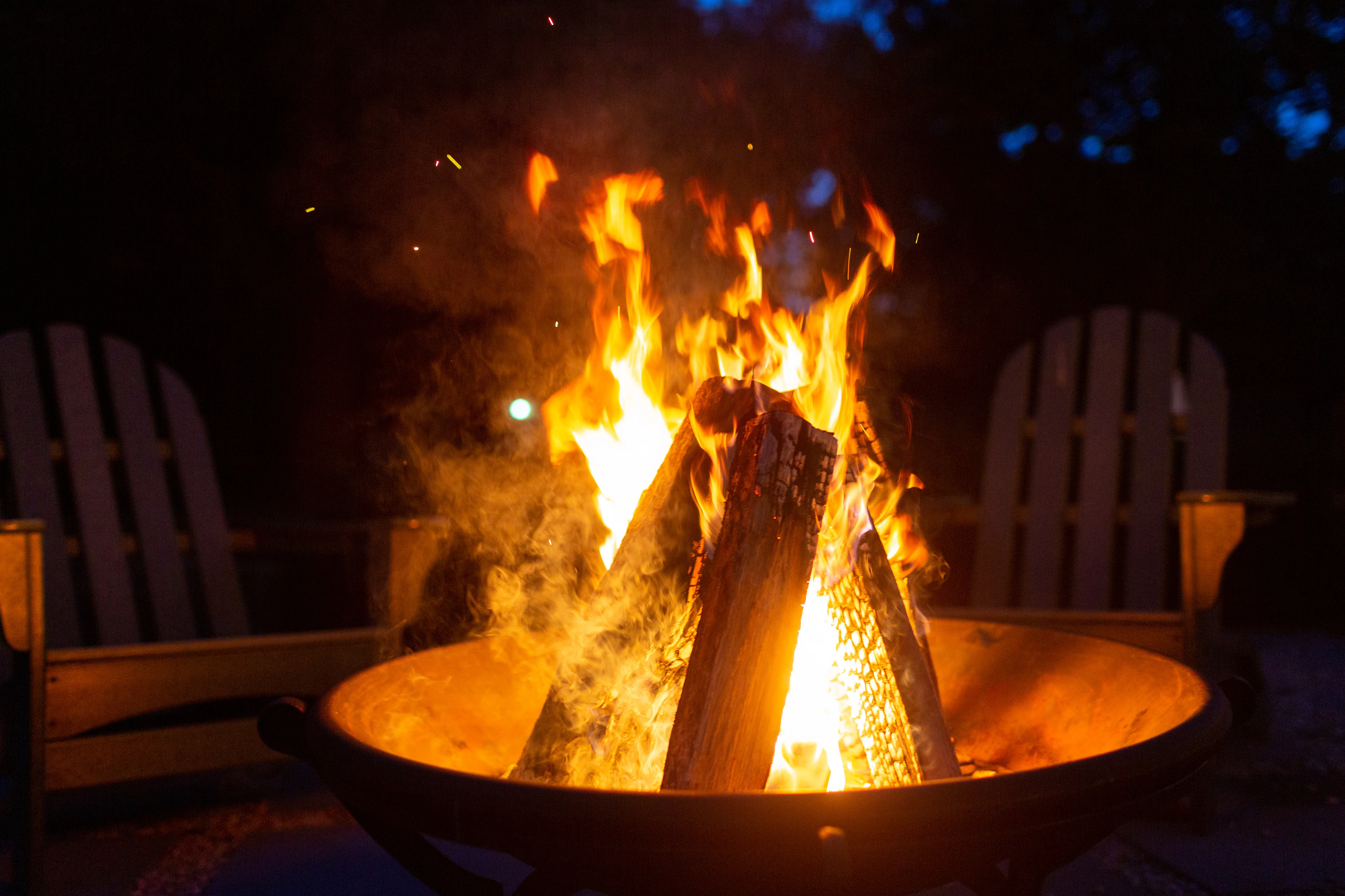 Wooden chairs surround a roaring fire pit which lets off and orange glow in the darkness of the night.