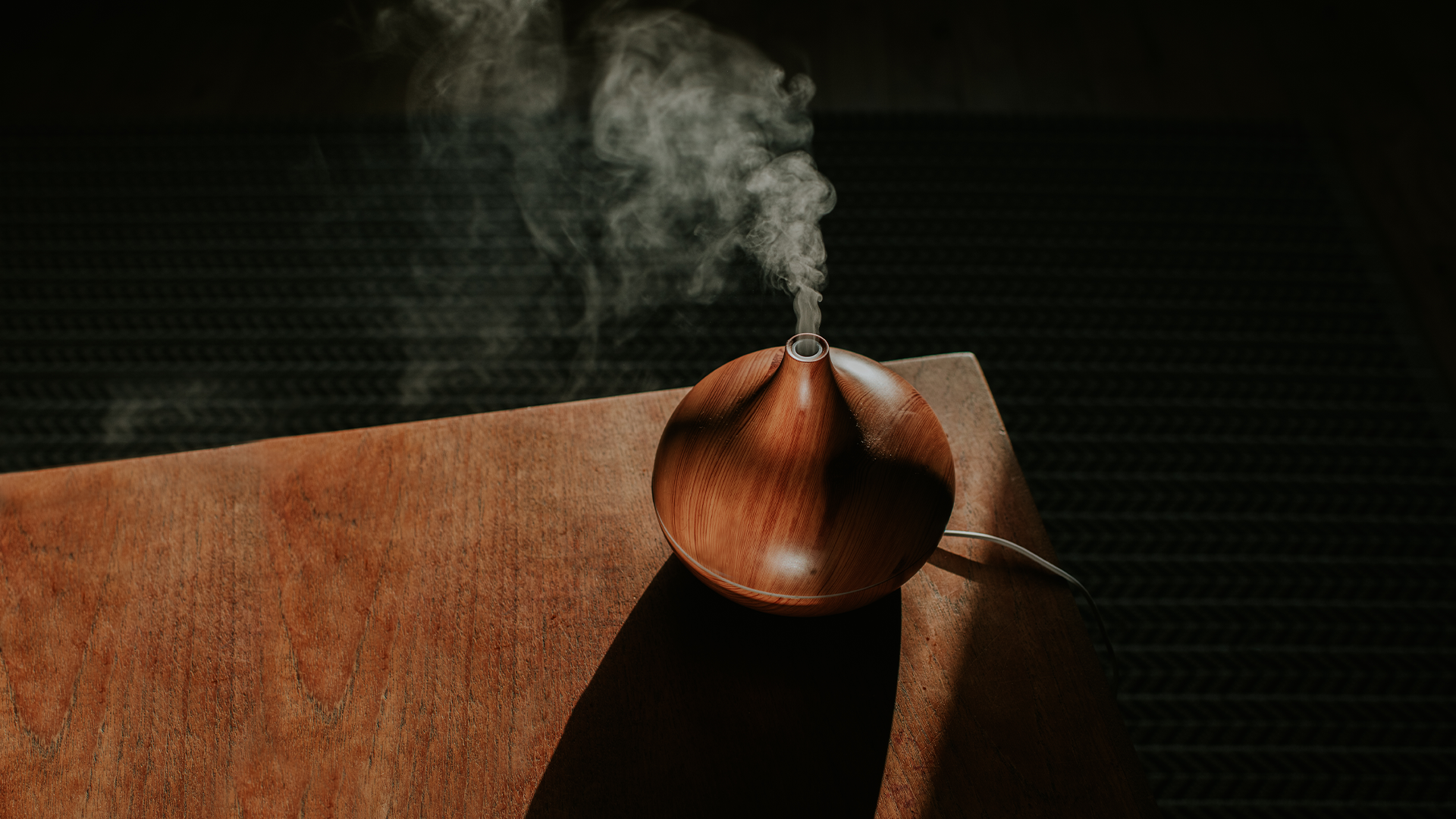 A wood-look aroma diffuser, placed on a wooden table in front of a black wall.
