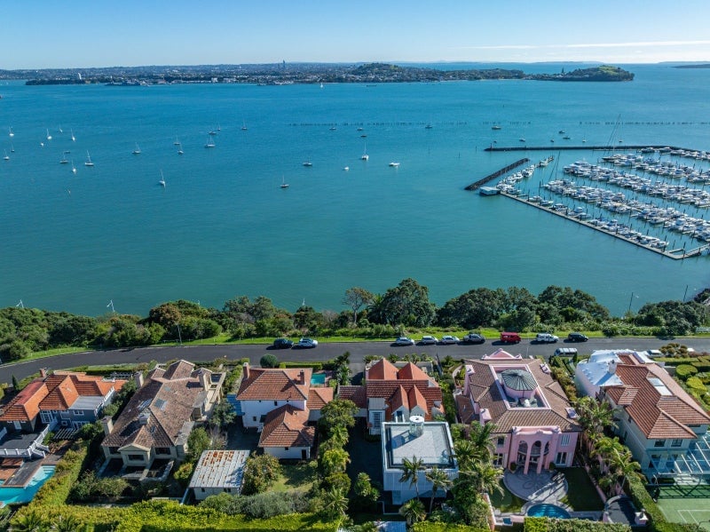 An aerial view of Paritai Drive's clifftop homes with red-tiled roofs lining the waterfront, their large sections separated by manicured hedges, overlooking the bright turquoise water and moored boats below