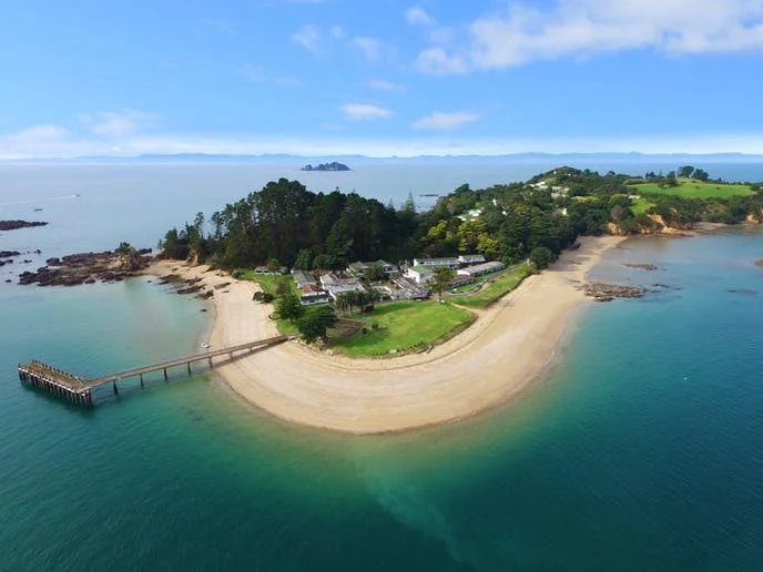 Aerial view of Pakatoa Island, one of New Zealand’s most expensive houses and private resorts, featuring golden sandy beaches, lush greenery, and a long pier stretching into the sea.