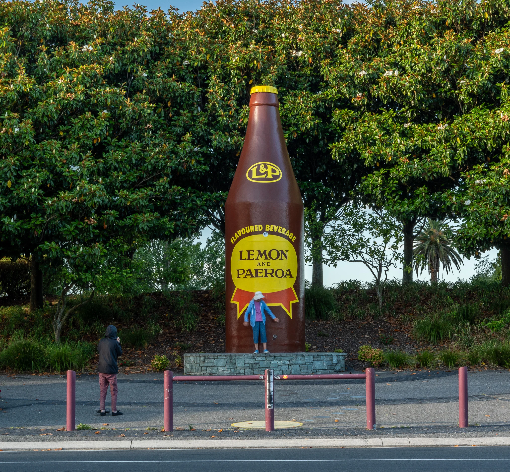 A large bottle stands tall amongst some trees. The label reads L&P which is a New Zealand beverage
