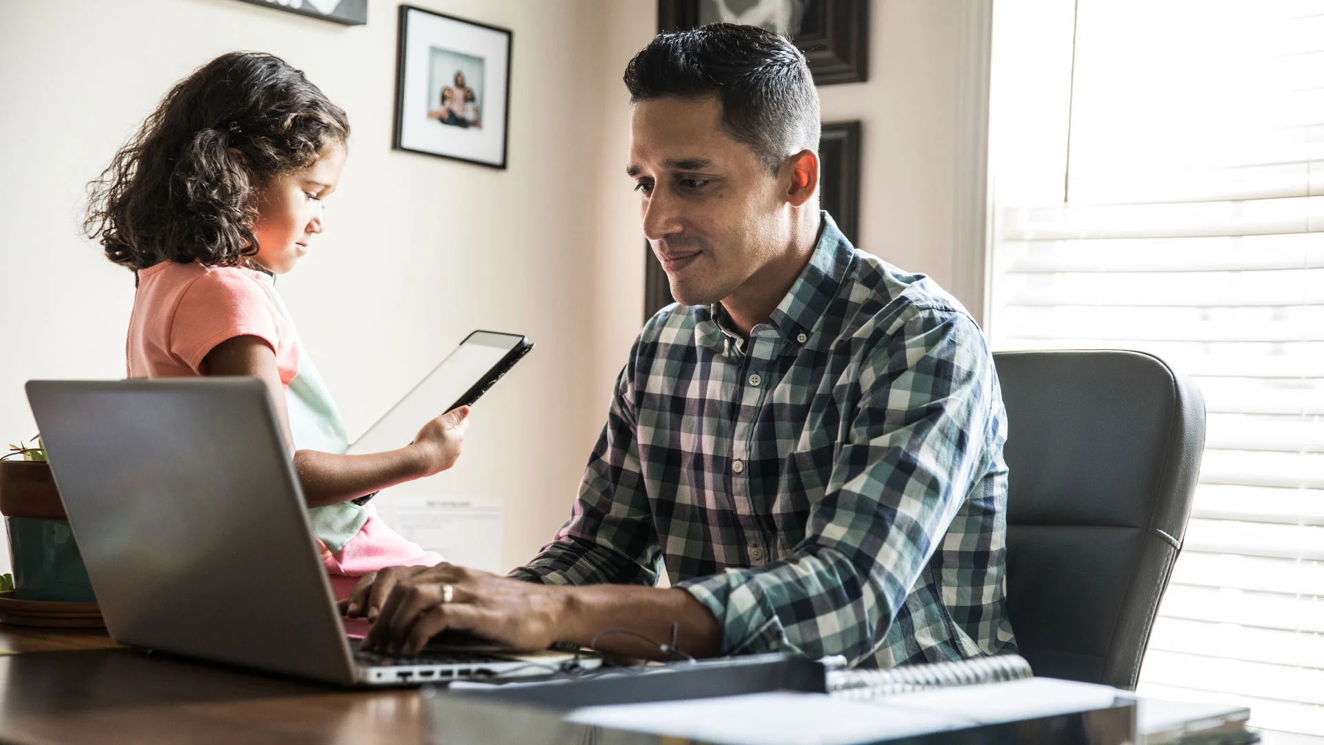 Young man working from home on a laptop while his young daughter plays on a tablet sitting next to him.