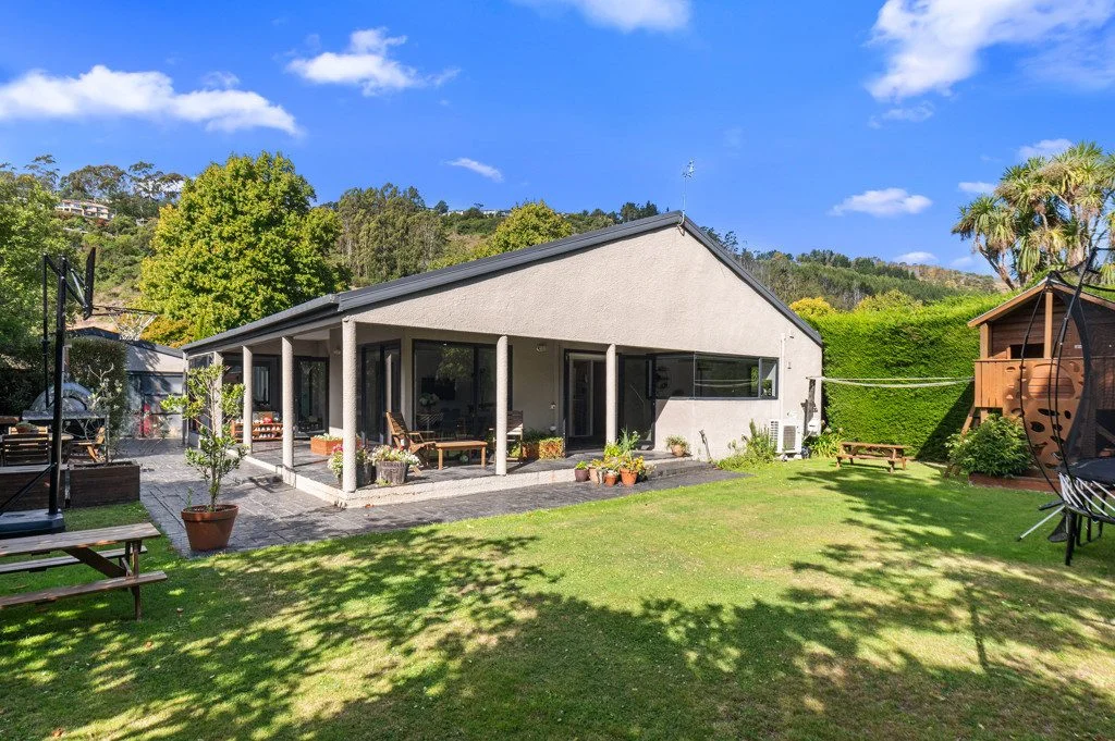 A classic Kiwi bungalow surrounded by lush green grass and manicured trees.