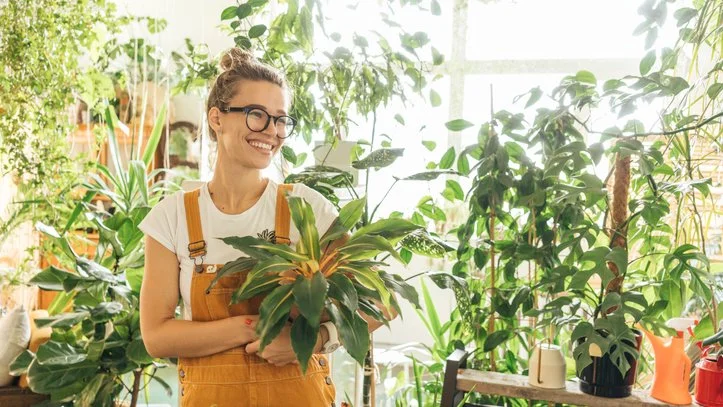 Woman standing in plant shop surrounded by hanging indoor plants. She is holding a peace lily.