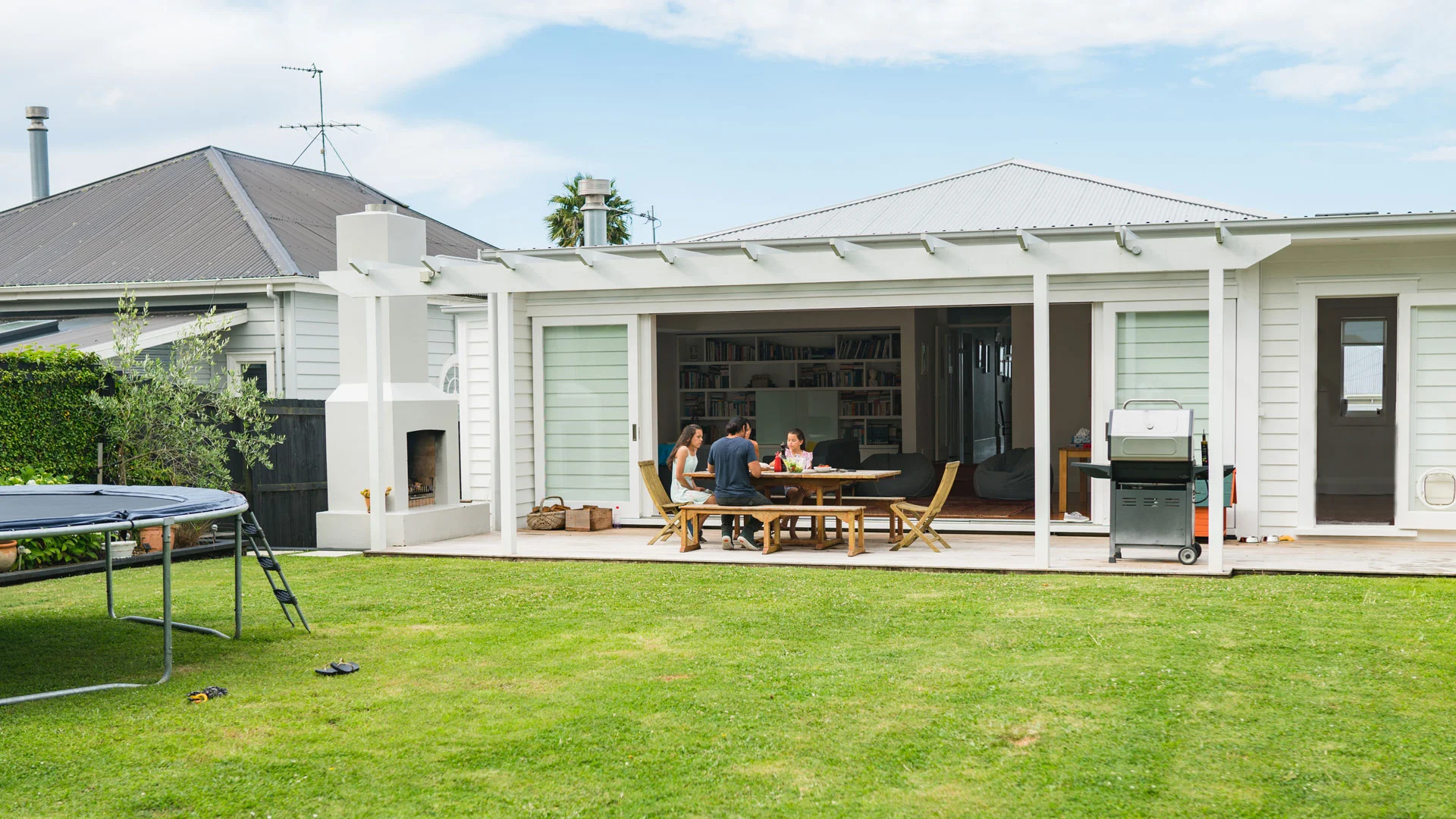 A NZ home with family sitting out on the deck having a meal, with a large green law nin the foreground.