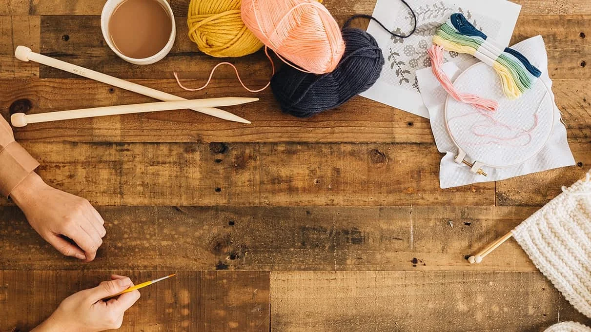 A table top full of craft items - yarn, knitting needles, embroidery supplies, a half knitted scarf, painting supplies and a cup of tea