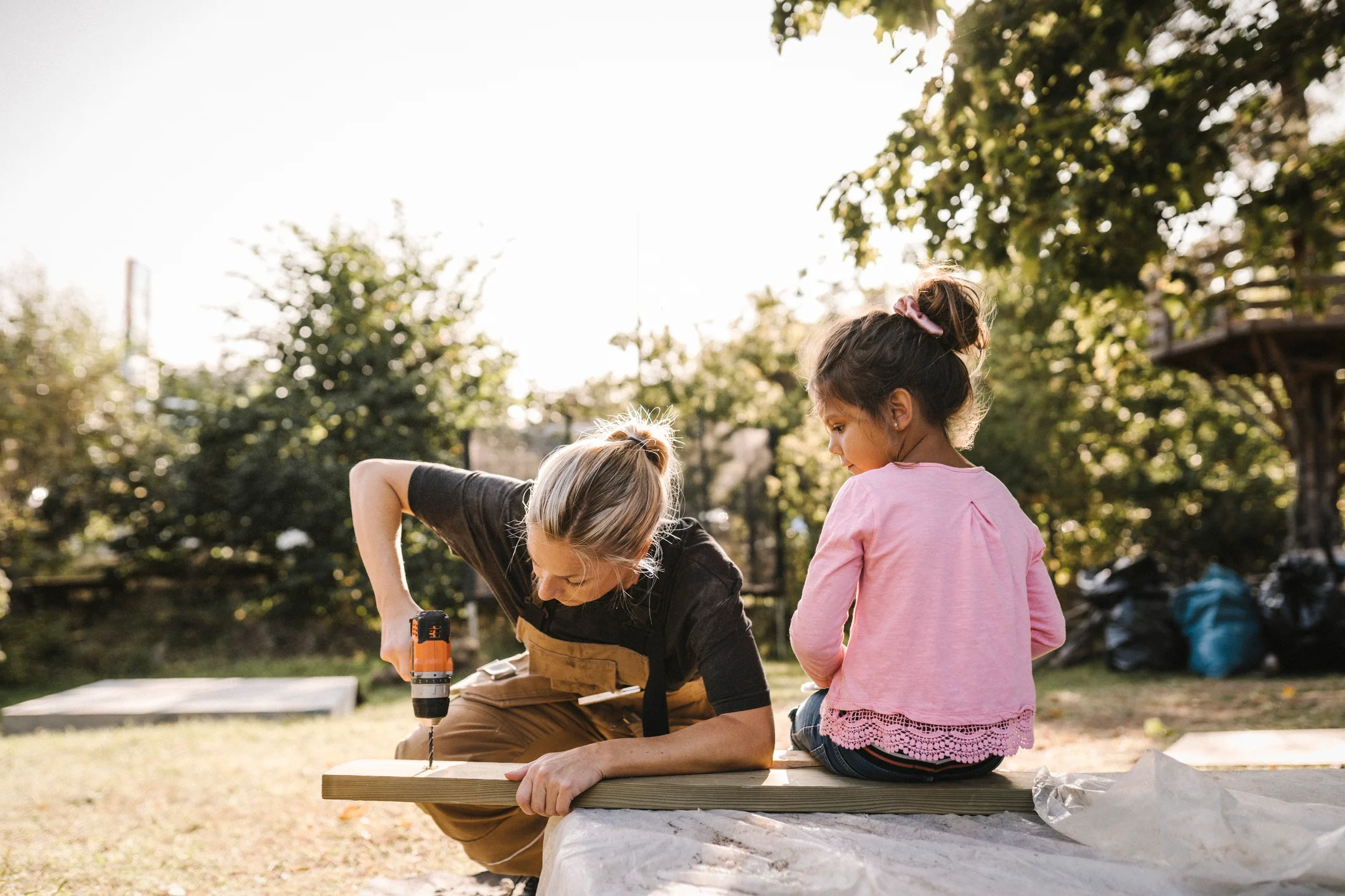 Mother uses a drill while daughter sits on plank of wood