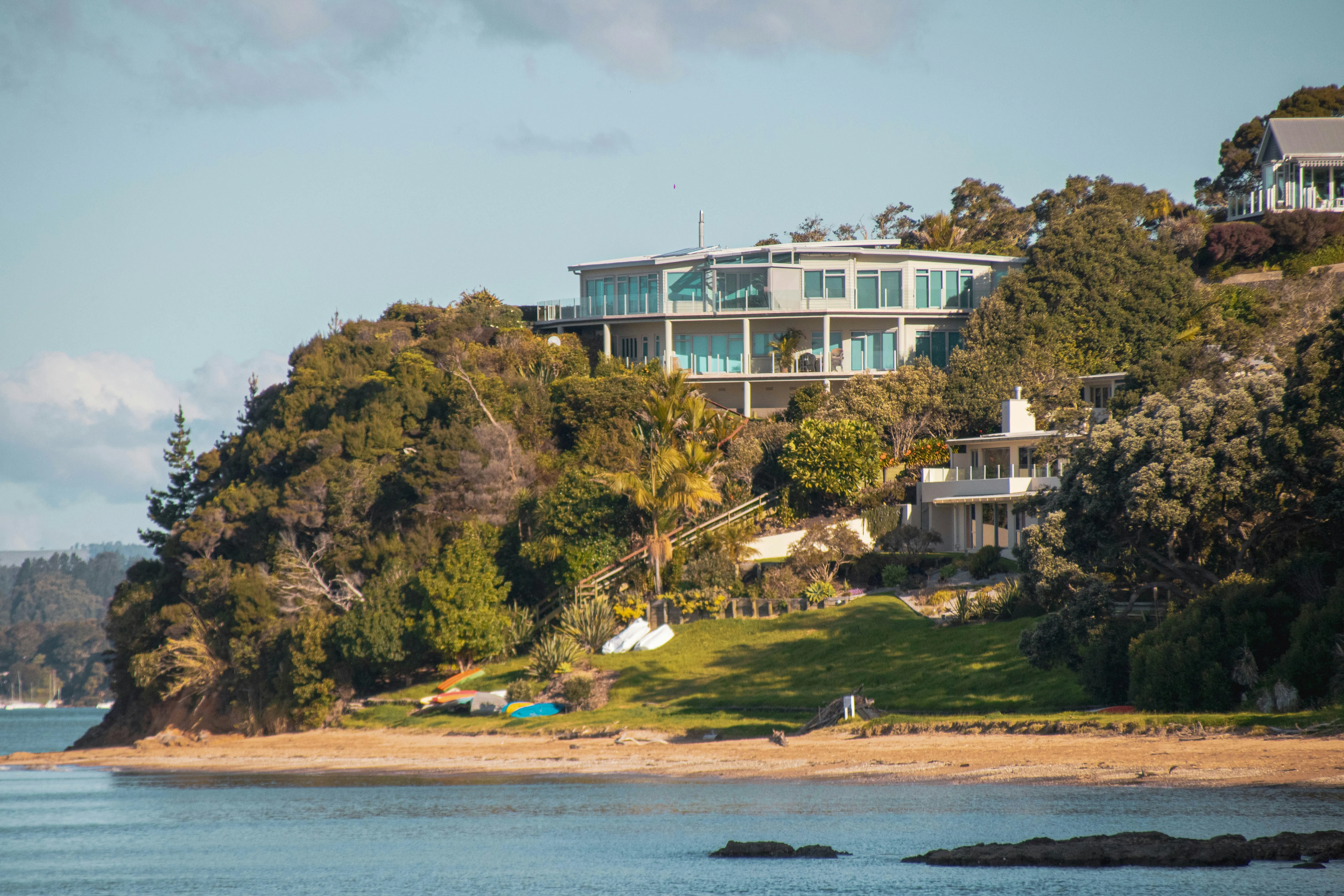 Modern glass-walled house perched on a hillside, overlooking a beach and calm waters below.