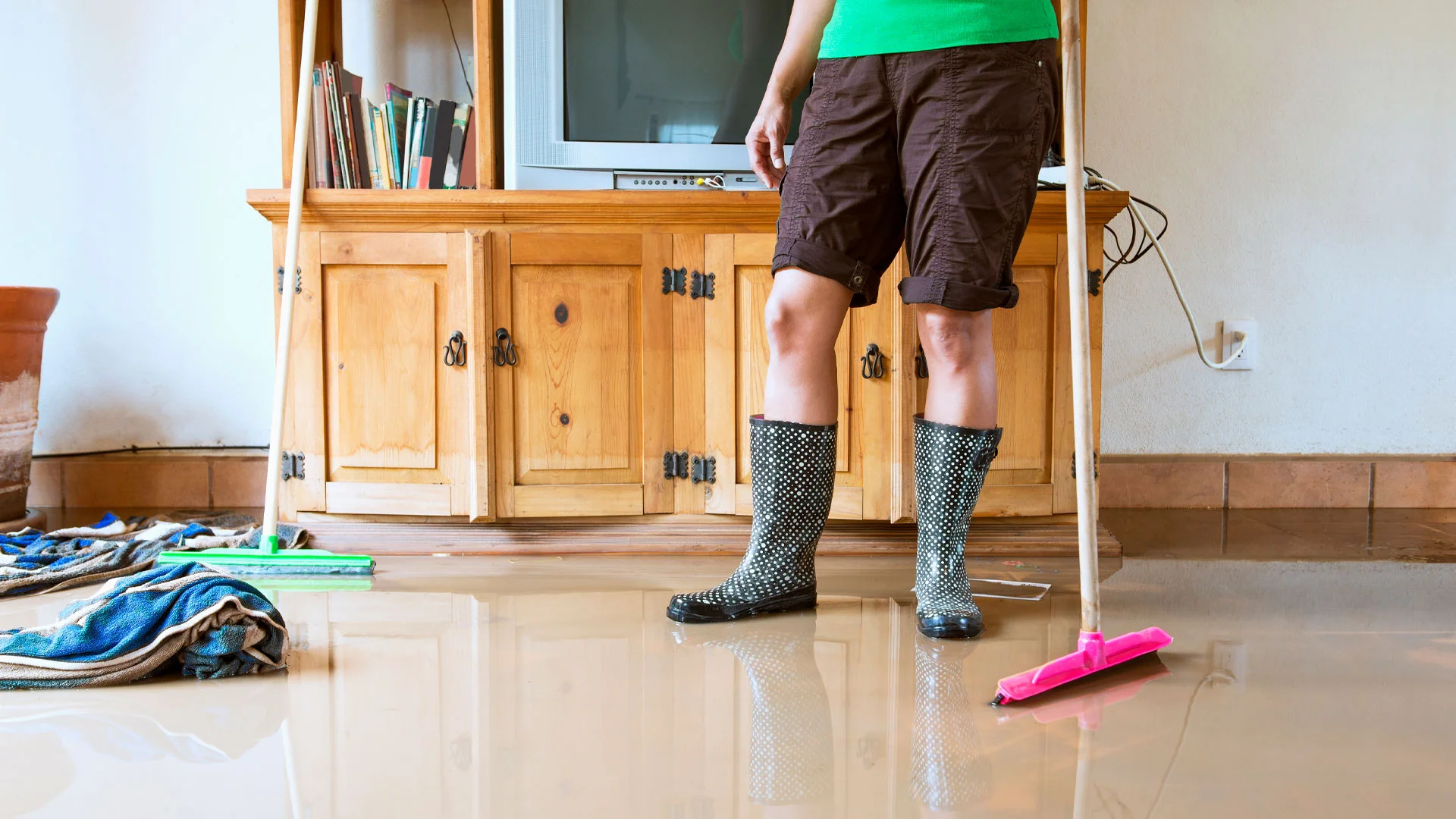Man standing in his living room in gumboots, with a mop, clearing up after a flood damaged his home in New Zealand.
