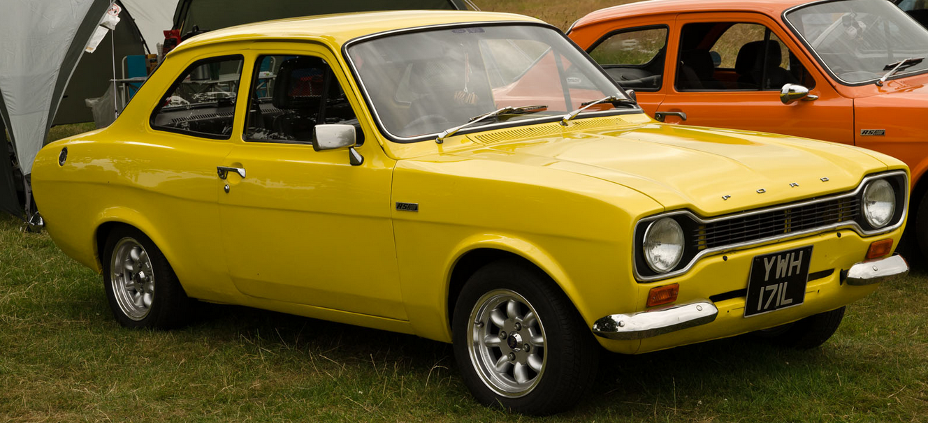Parked on grass in front of a tent: Old yellow ford escort in the foreground, partially visible is an orange Ford escort just behind it