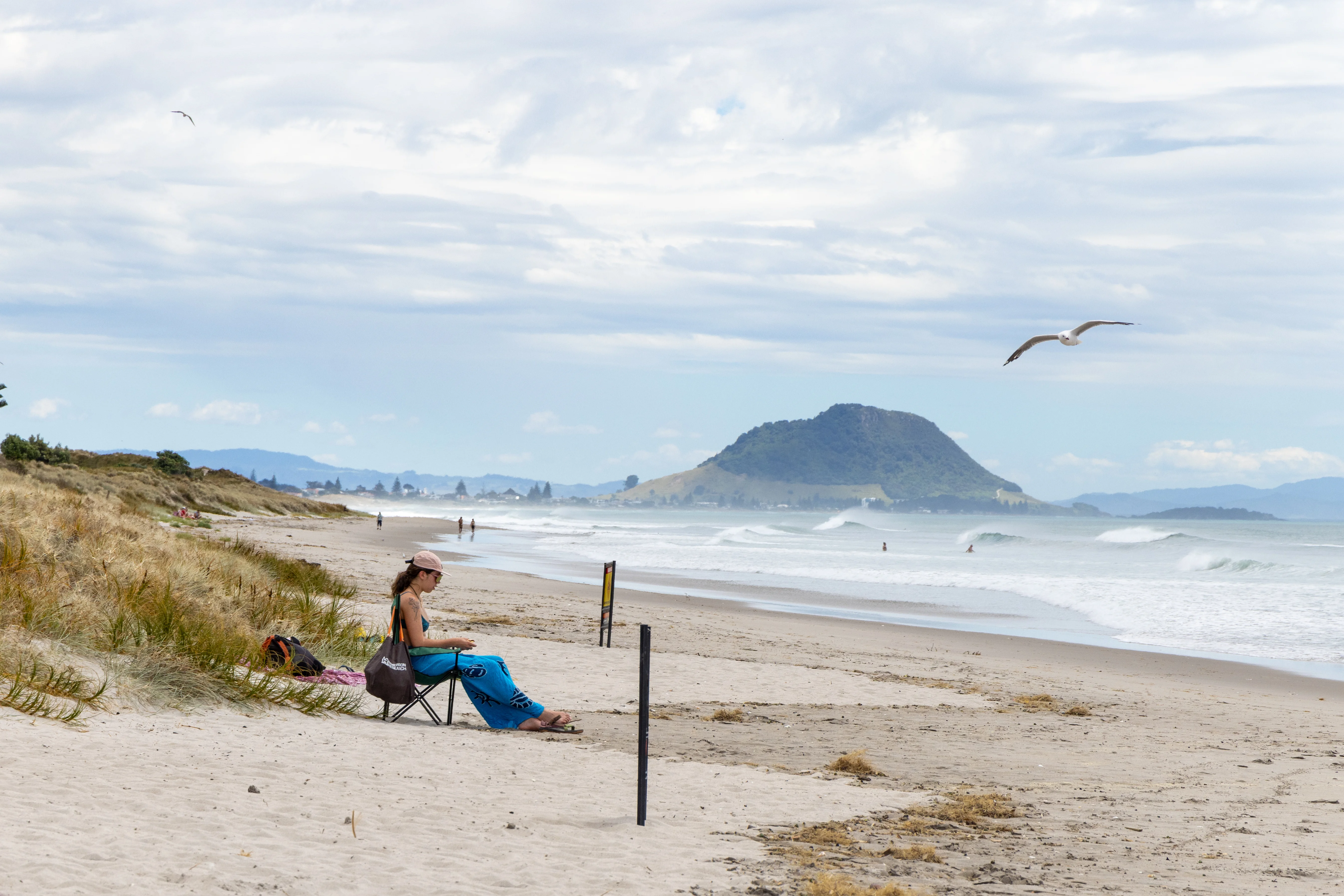 Golden beach with waves in the background. A woman sits on the sand dunes looking out to the sea. 
