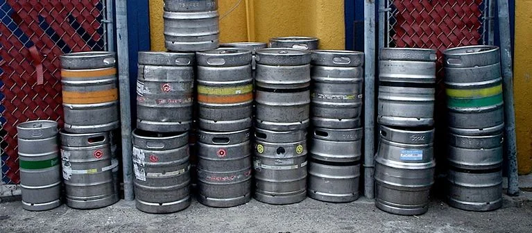 a line of aluminium beer kegs stacked up by a chain link fence with yellow doors behind the kegs